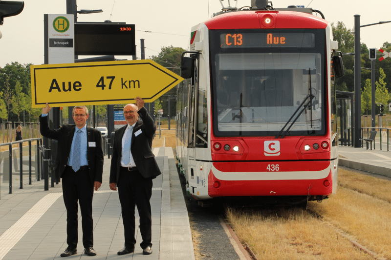 Citylink im Haltepunkt Technopark. Ein Pfeilförmiges Hinweisschild mit der Beschriftung "Aue 47 km" wird von zwei Männern in Anzügen lächelnd hochgehalten