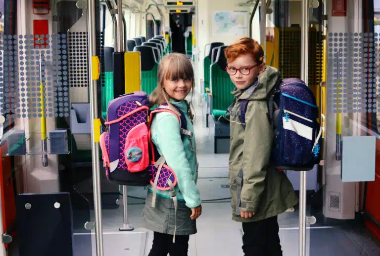 Zwei Kinder im Gang (Bereich der Türen) eines Citylinks, beiden haben ihren Schulranzen auf dem Rücken, das Mädchen links lächelt in die Kamera, der Junge rechts trägt eine Brille und schmunzelt