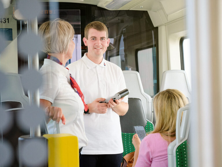 In einem Citylink: Eine Kundenbetreuerin begleitet einen jungen Mann, dieser lächelt in die Kamera, vor den beiden sitzt eine Kundin und zeigt ihr Smartphone mit dem digitalen Ticket vor