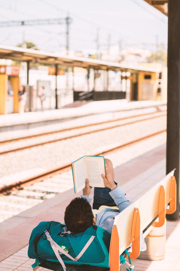 Ein junger Mann liegt an einem sonnigen Tag auf einer Bank am Bahnsteig und liest ein Buch. Dabei ruht sein Kopf auf seiner Tasche.